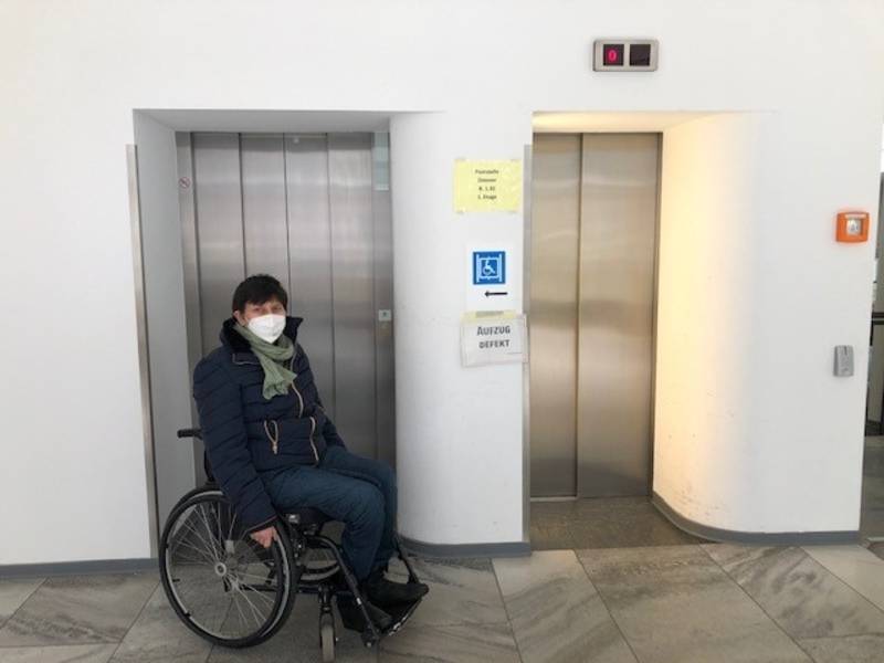 Frau im Rollstuhl vor einem defekten Aufzug